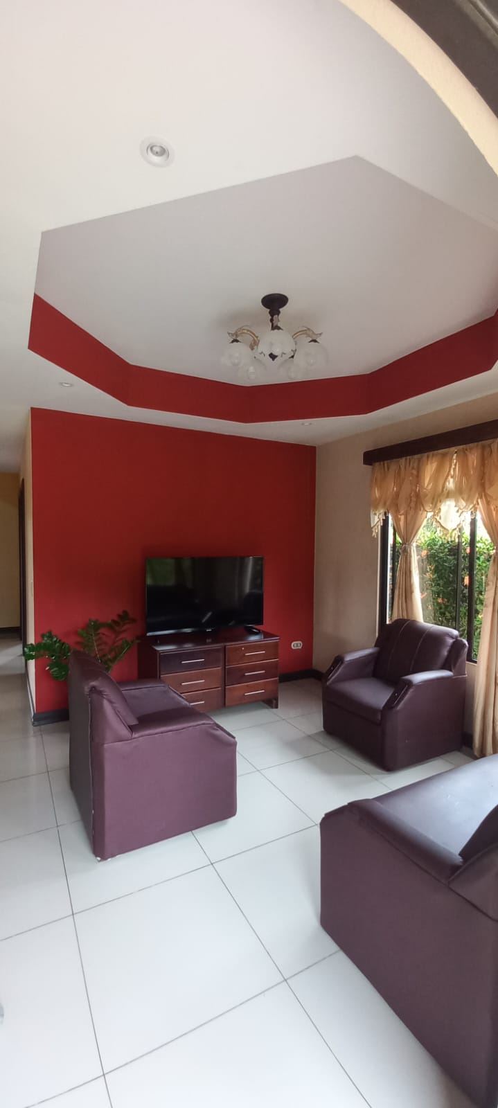 Living room with a red accent wall, a flat-screen TV on a wooden console, and burgundy leather chairs by a large window with beige curtains.