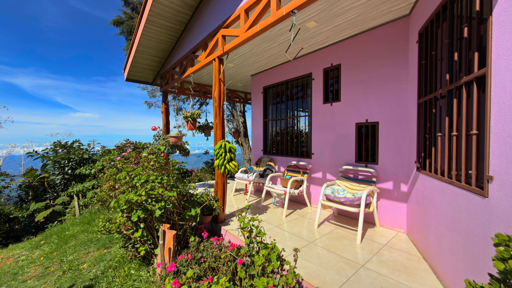 Pink-walled porch with brown barred windows, plastic chairs, and hanging plants against a hillside and blue sky garden view.