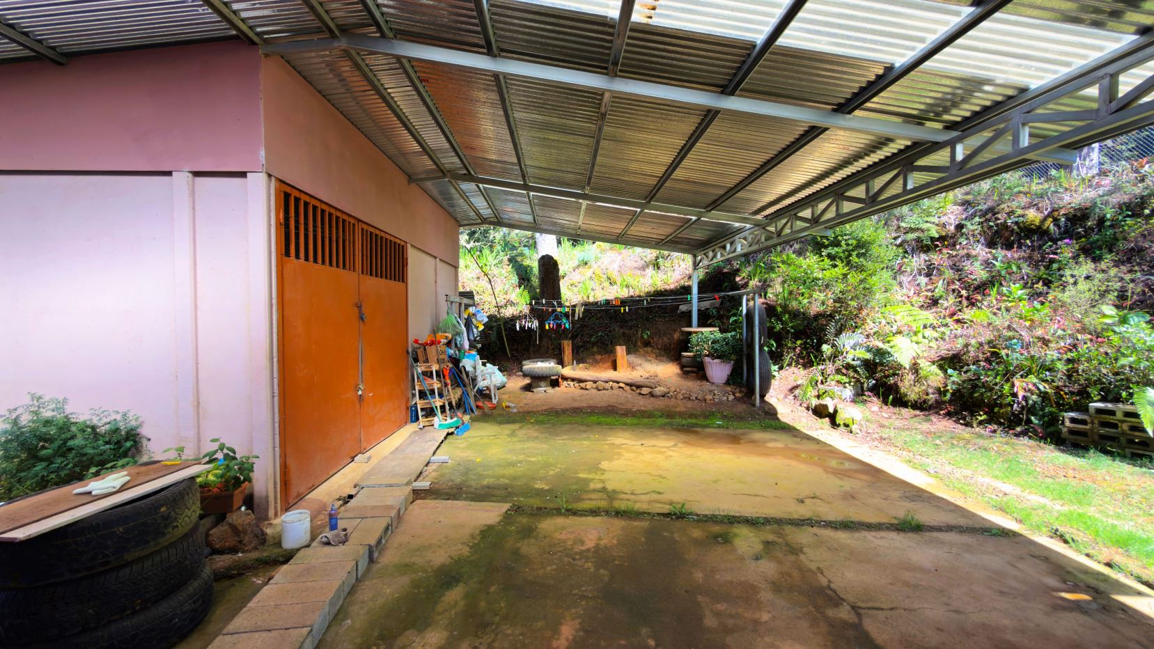 Covered outdoor workspace attached to a pink wall with orange double doors, cluttered with tools and plants under a translucent metal roof.