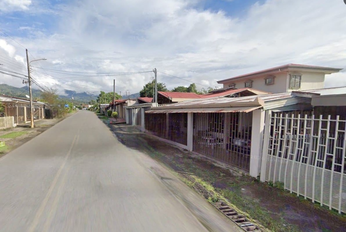 Residential street with single-story houses and metal fences on the right, extends toward distant hills under a partly cloudy sky.
