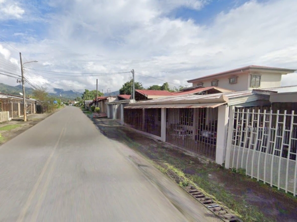 Residential street with single-story houses and metal fences on the right, extends toward distant hills under a partly cloudy sky.