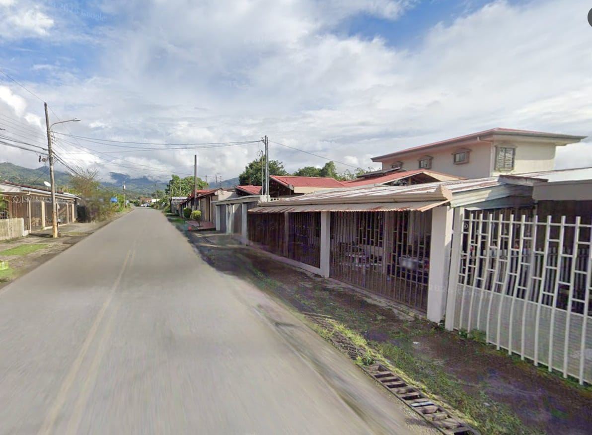 Residential street with single-story houses and metal fences on the right, extends toward distant hills under a partly cloudy sky.
