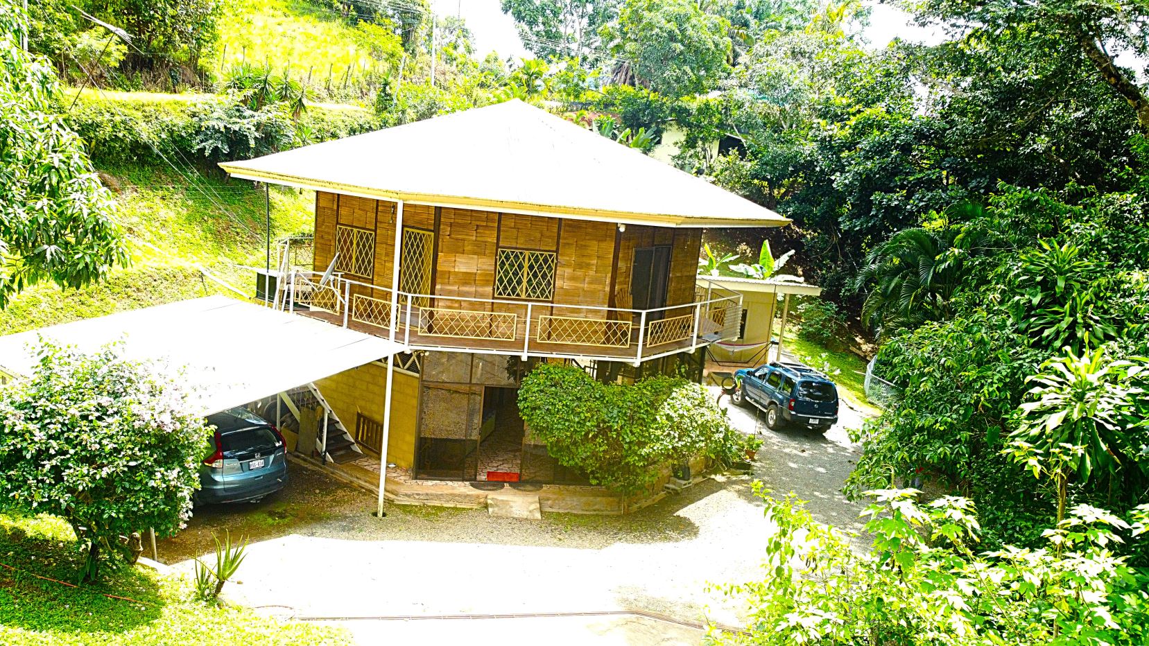 Two-story bamboo-clad house with a wraparound white railing, set on a grassy hillside with tropical trees and two parked cars nearby.