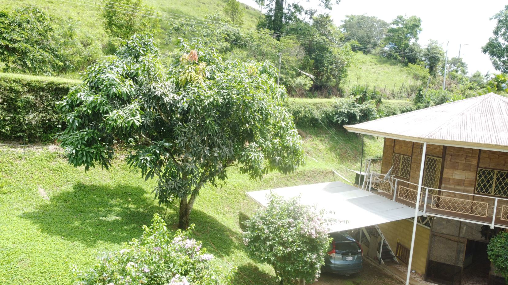 Wooden house with a light metal roof on a slope, balcony railing visible, car parked under a covered area amid green lawn and trees.