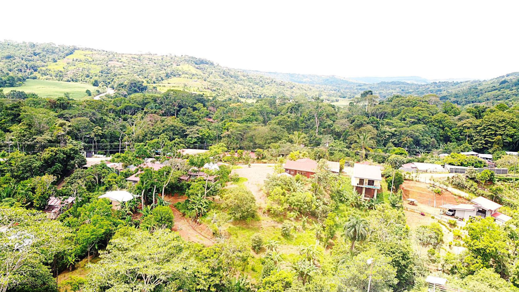 Aerial view of a rural village surrounded by dense jungle and rolling hills beneath a bright sky and distant mountains.