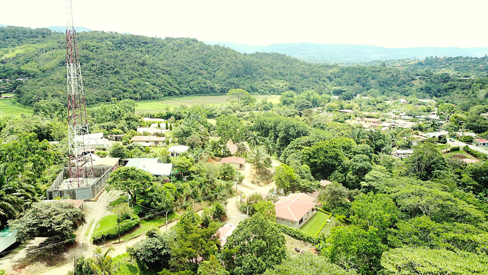 Aerial view of a rural village with dense greenery and hills, featuring a tall red-and-white communications tower in the foreground.