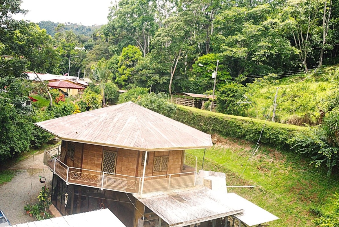 Wooden house with a wraparound balcony on a grassy hillside, surrounded by dense tropical trees.