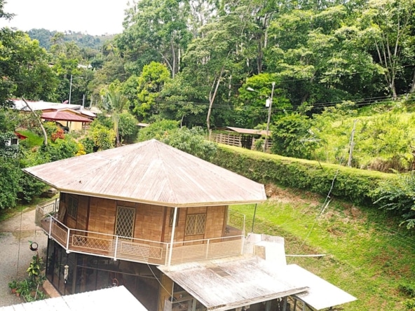 Wooden house with a wraparound balcony on a grassy hillside, surrounded by dense tropical trees.