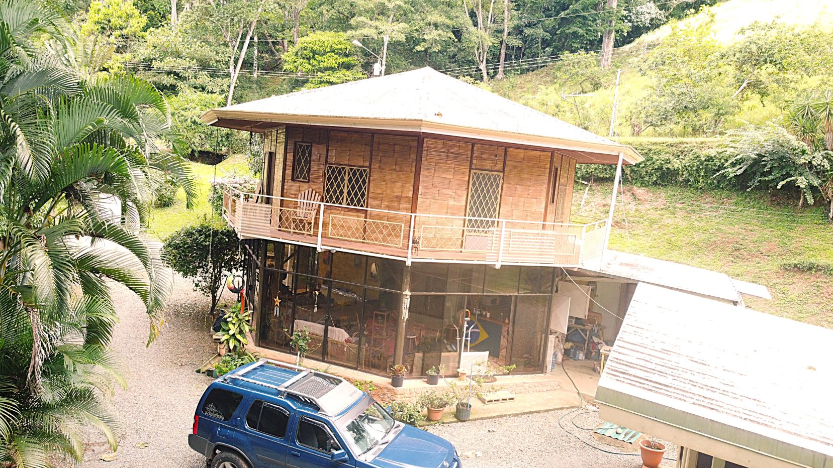 Two-story wooden house on stilts with a glass-front ground level surrounded by plants and a blue SUV parked nearby.