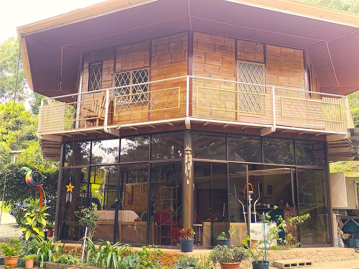 Two-story bamboo-clad house with a glass storefront on the ground floor, and a wooden balcony with lattice rails above, surrounded by potted plants.