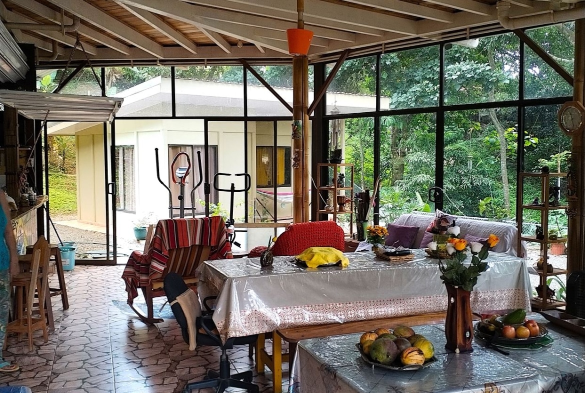 Bright indoor garden room with glass walls, a long table covered in plastic, fruit bowls and flowers, and exercise equipment in the back yard view outside to greenery.