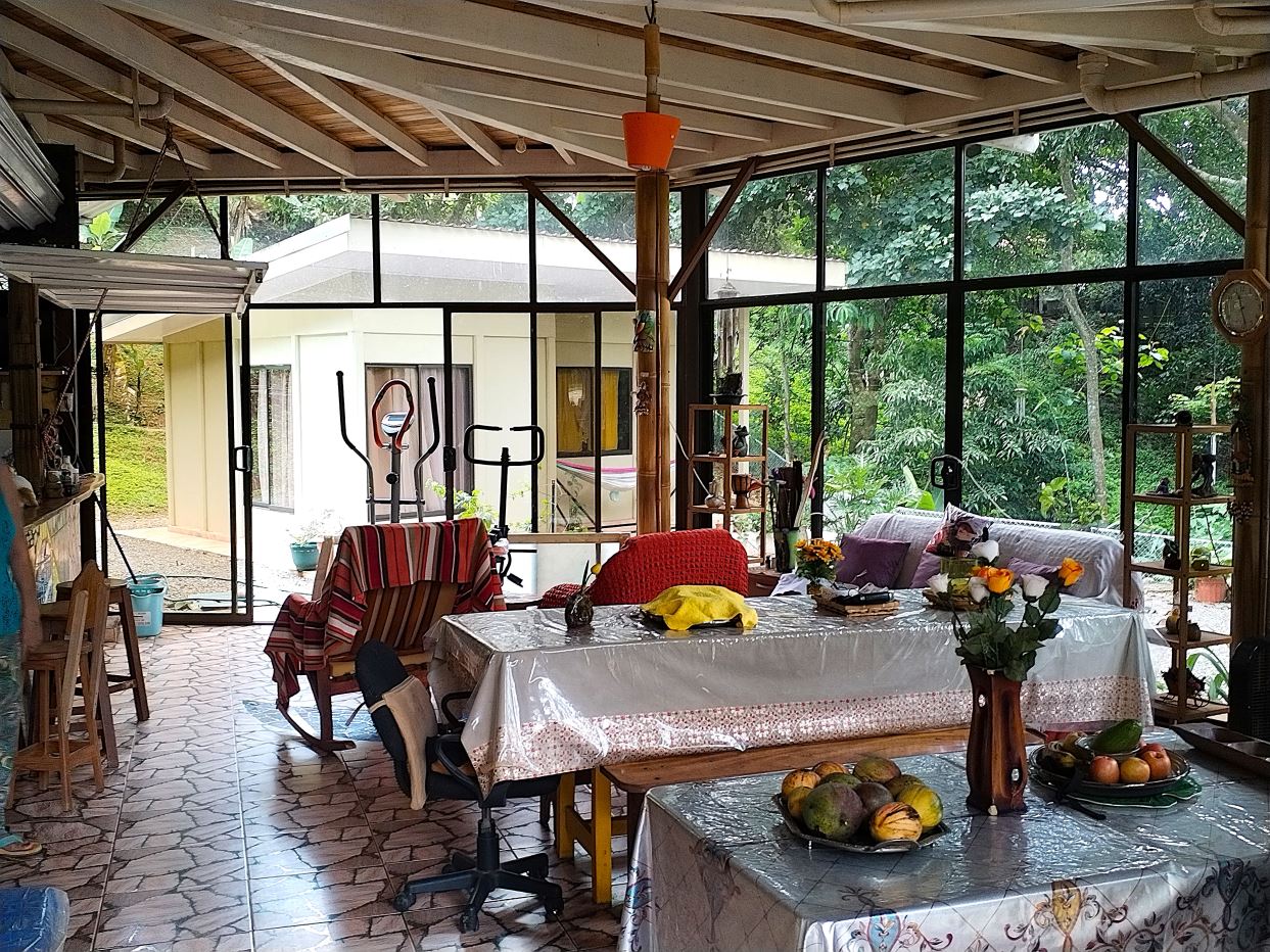 Bright indoor garden room with glass walls, a long table covered in plastic, fruit bowls and flowers, and exercise equipment in the back yard view outside to greenery.