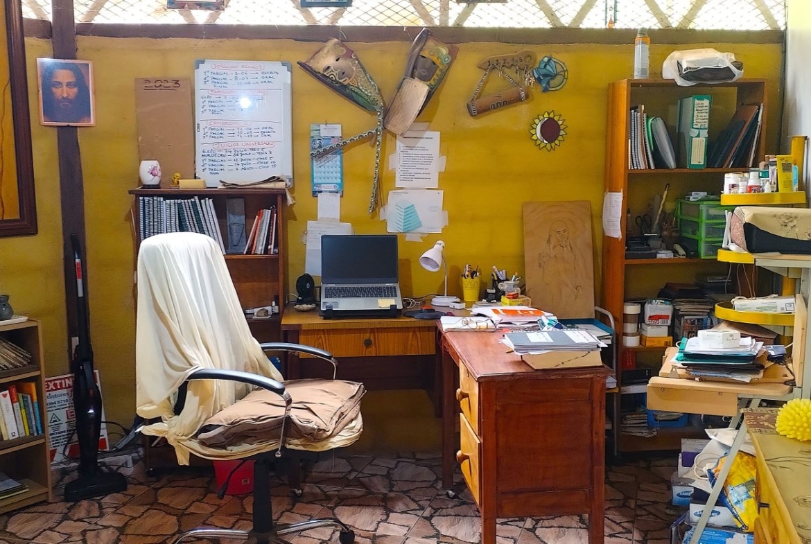 Home office cluttered desk with laptop, lamp, papers, and a chair draped in a white cloth.
