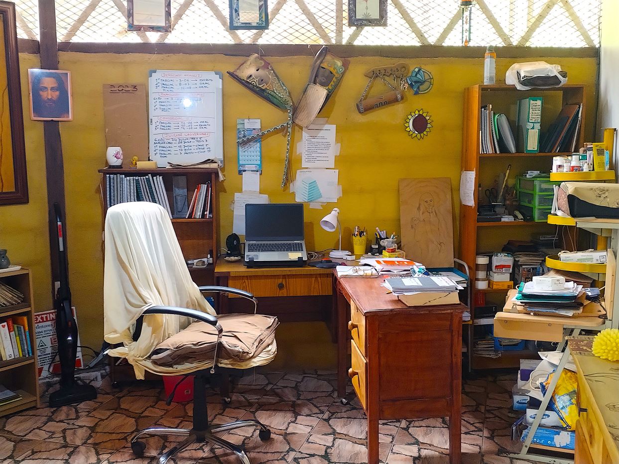 Home office cluttered desk with laptop, lamp, papers, and a chair draped in a white cloth.