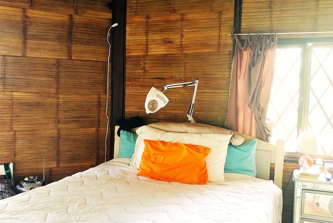 Bedroom with bamboo walls, white quilted bed, orange accent pillow and teal pillows; a white adjustable lamp above the headboard creates a cozy reading nook.