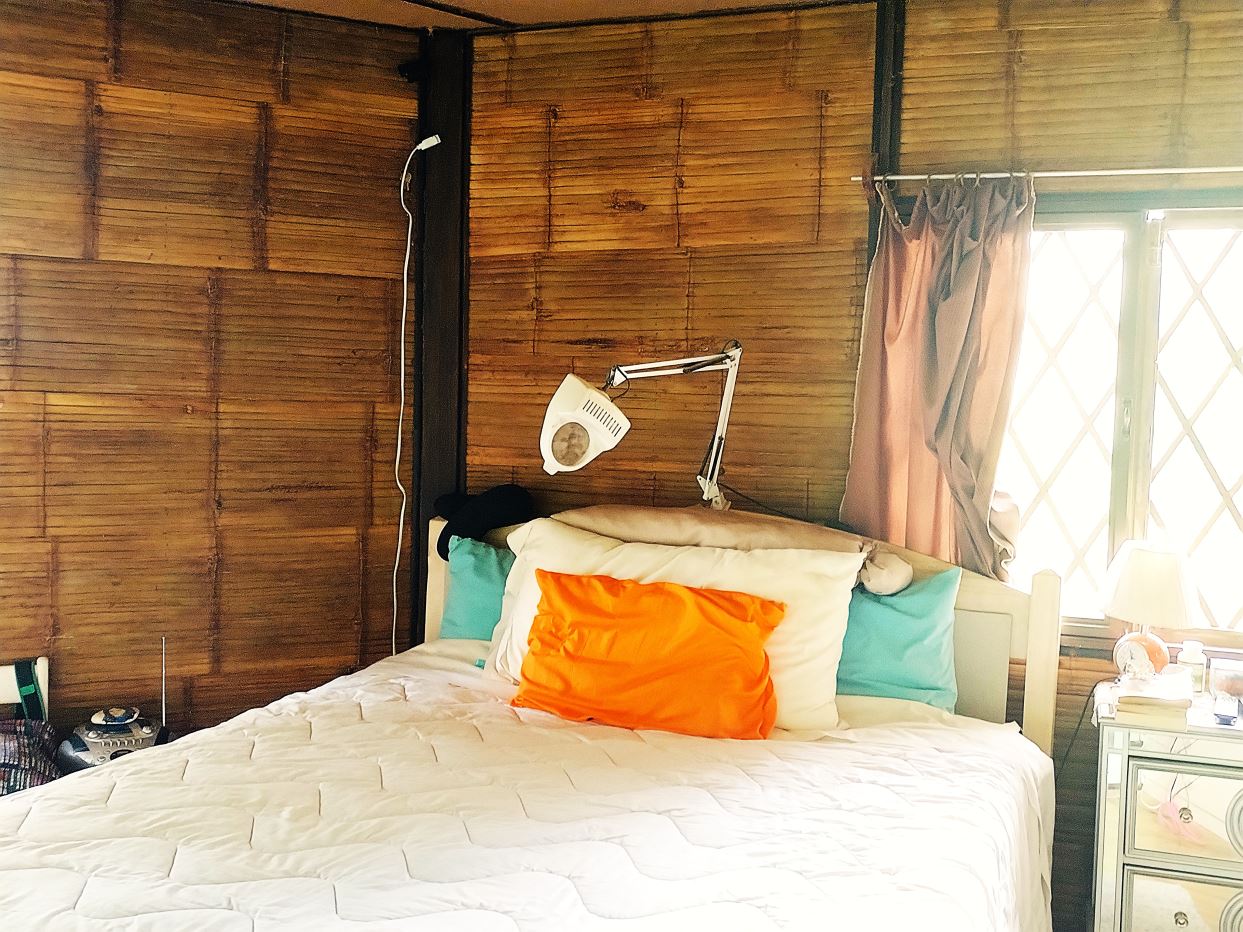 Bedroom with bamboo walls, white quilted bed, orange accent pillow and teal pillows; a white adjustable lamp above the headboard creates a cozy reading nook.