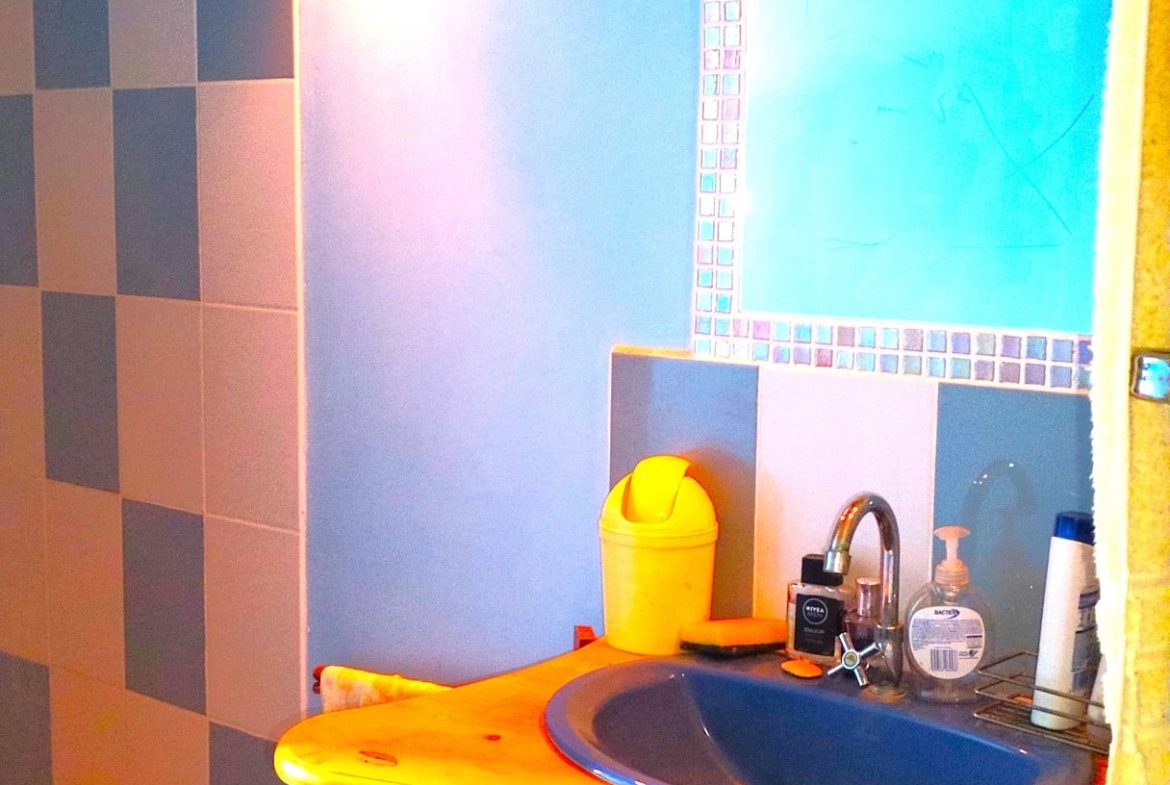 Bright blue bathroom with mosaic-framed mirror above a yellow sink cabinet, yellow disposal bin, and assorted toiletries on the counter