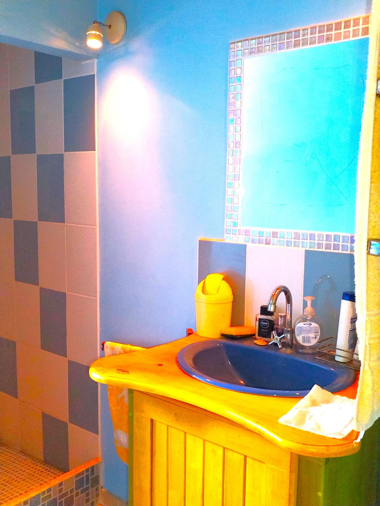 Bright blue bathroom with mosaic-framed mirror above a yellow sink cabinet, yellow disposal bin, and assorted toiletries on the counter