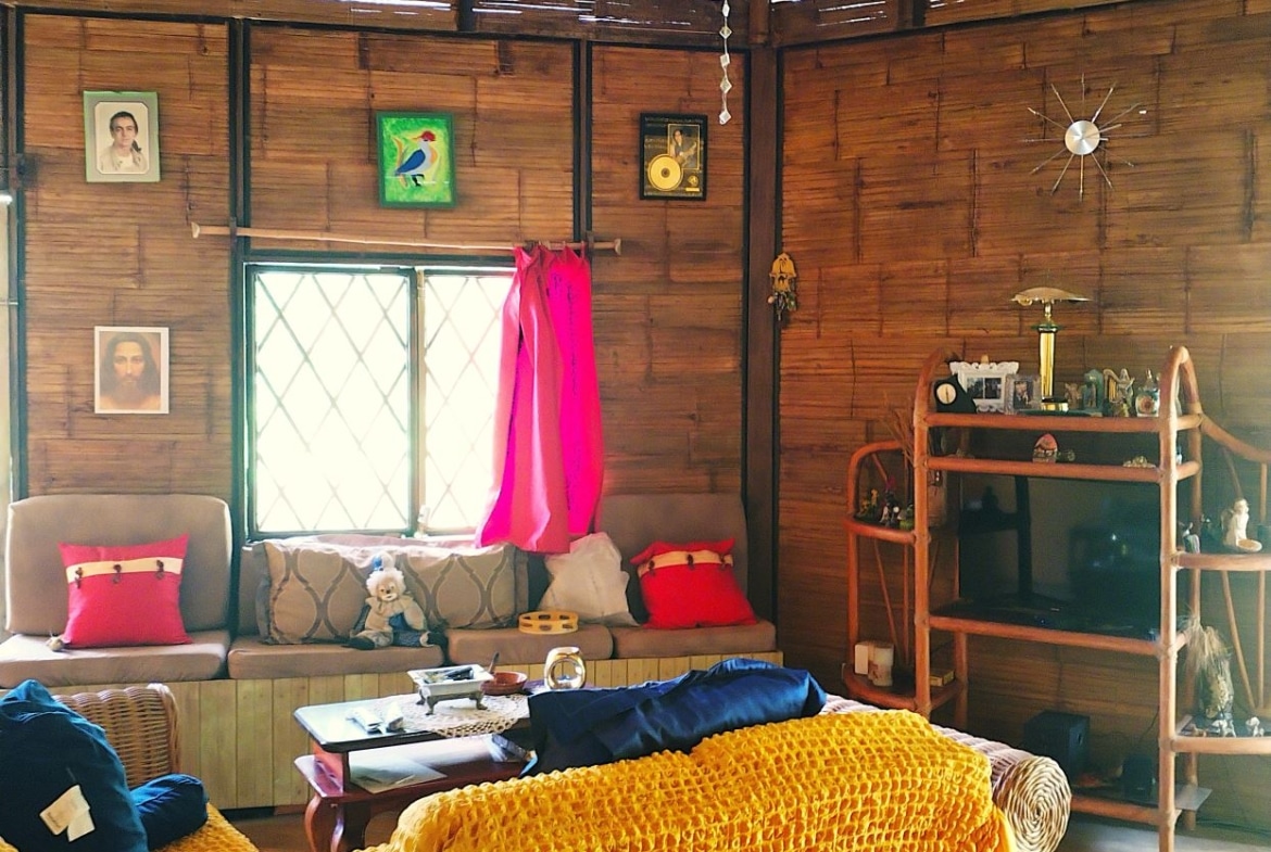 A cozy living room with rattan walls, a beige sofa, pink cushions, and a bright yellow crocheted throw over the armchair near a window with a pink curtain.