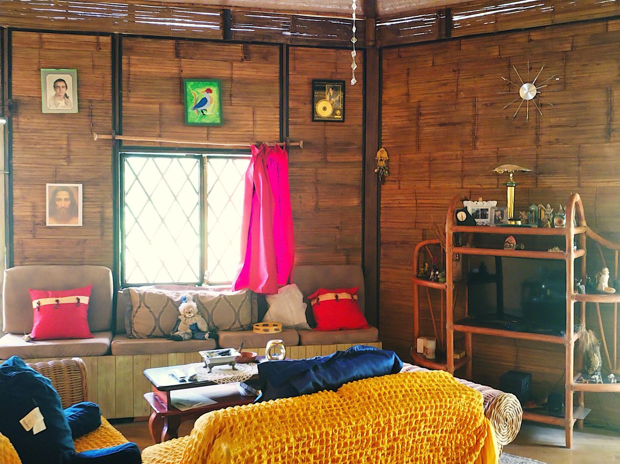A cozy living room with rattan walls, a beige sofa, pink cushions, and a bright yellow crocheted throw over the armchair near a window with a pink curtain.