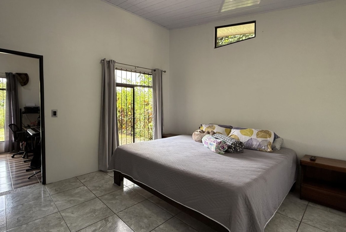 Bedroom with a large bed, gray bedspread and patterned pillows, two wooden nightstands, and a doorway leading to a small home office area visible to the left.