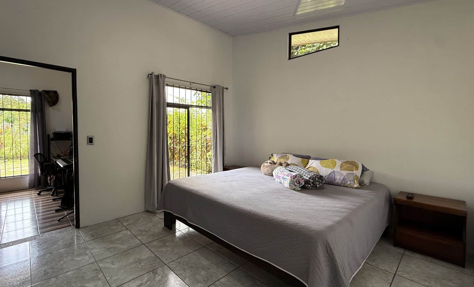 Bedroom with a large bed, gray bedspread and patterned pillows, two wooden nightstands, and a doorway leading to a small home office area visible to the left.
