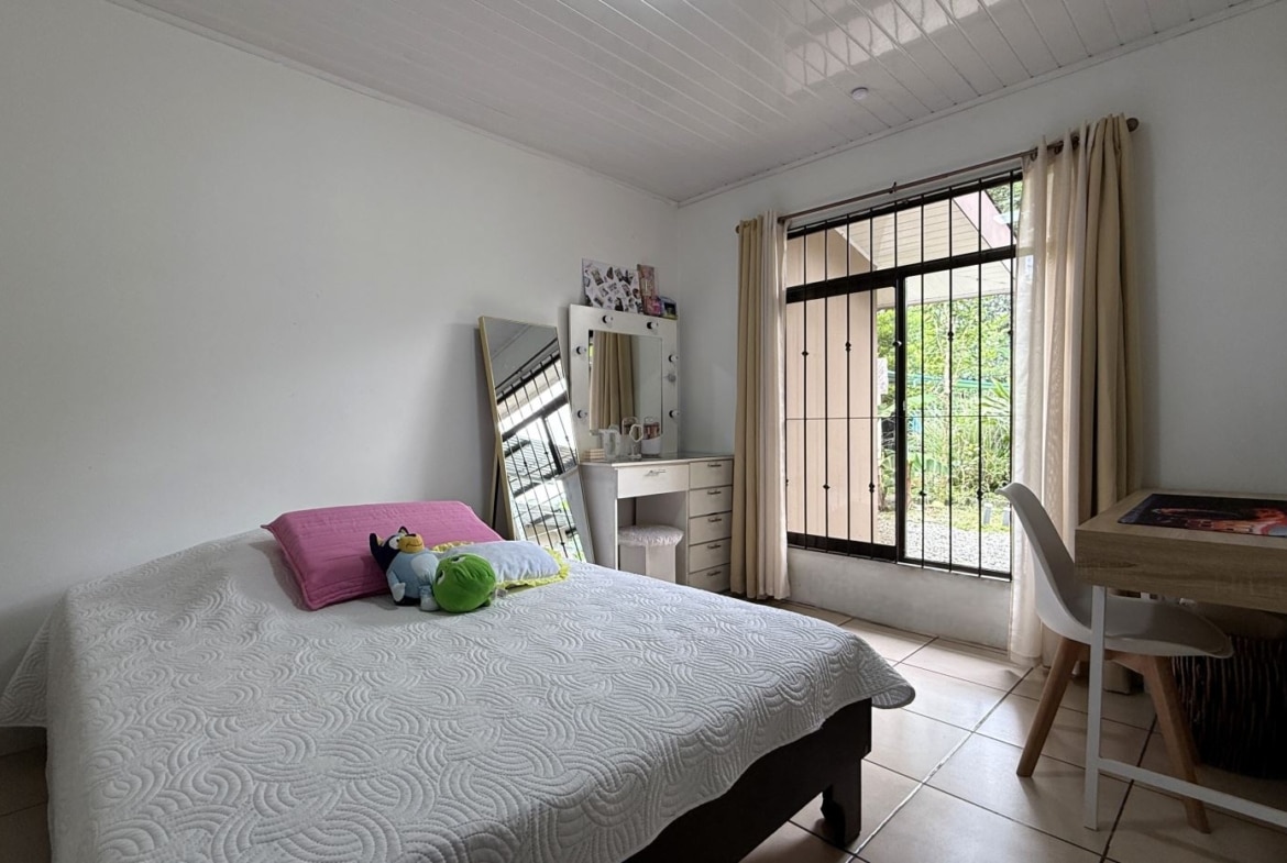 A simple bedroom with a bed, pink pillow, and plush toys, facing a vanity with a lit mirror and a leaning mirror nearby.