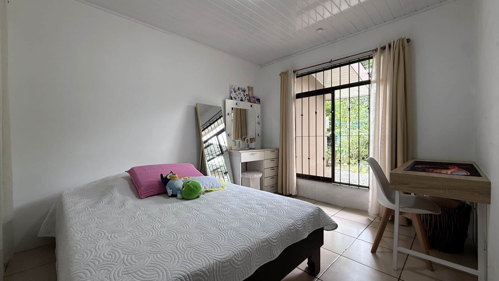 A simple bedroom with a bed, pink pillow, and plush toys, facing a vanity with a lit mirror and a leaning mirror nearby.