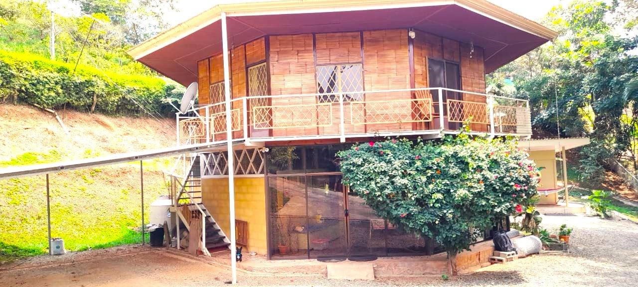 Two-story wooden house on a hillside with a wraparound balcony and a flowering shrub in front garden.