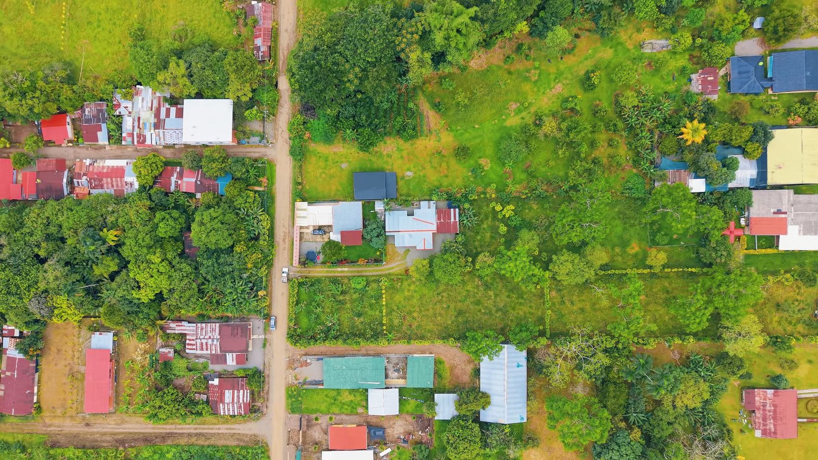Aerial view of a village with red-roofed houses along dirt streets amid dense green trees and open grassy plots.