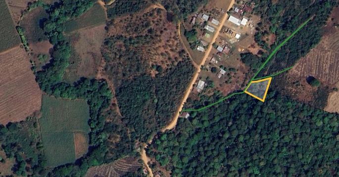 Aerial view of a rural area with fields and a small village; a yellow-outlined triangular plot sits beside a road near dense forest.