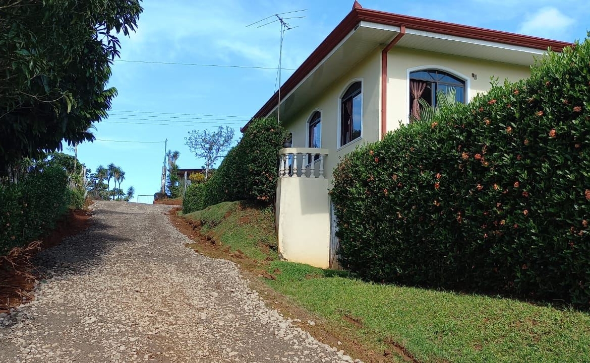 Gravel road slopes uphill beside a yellow house with arched windows, bordered by tall hedges under a blue sky.