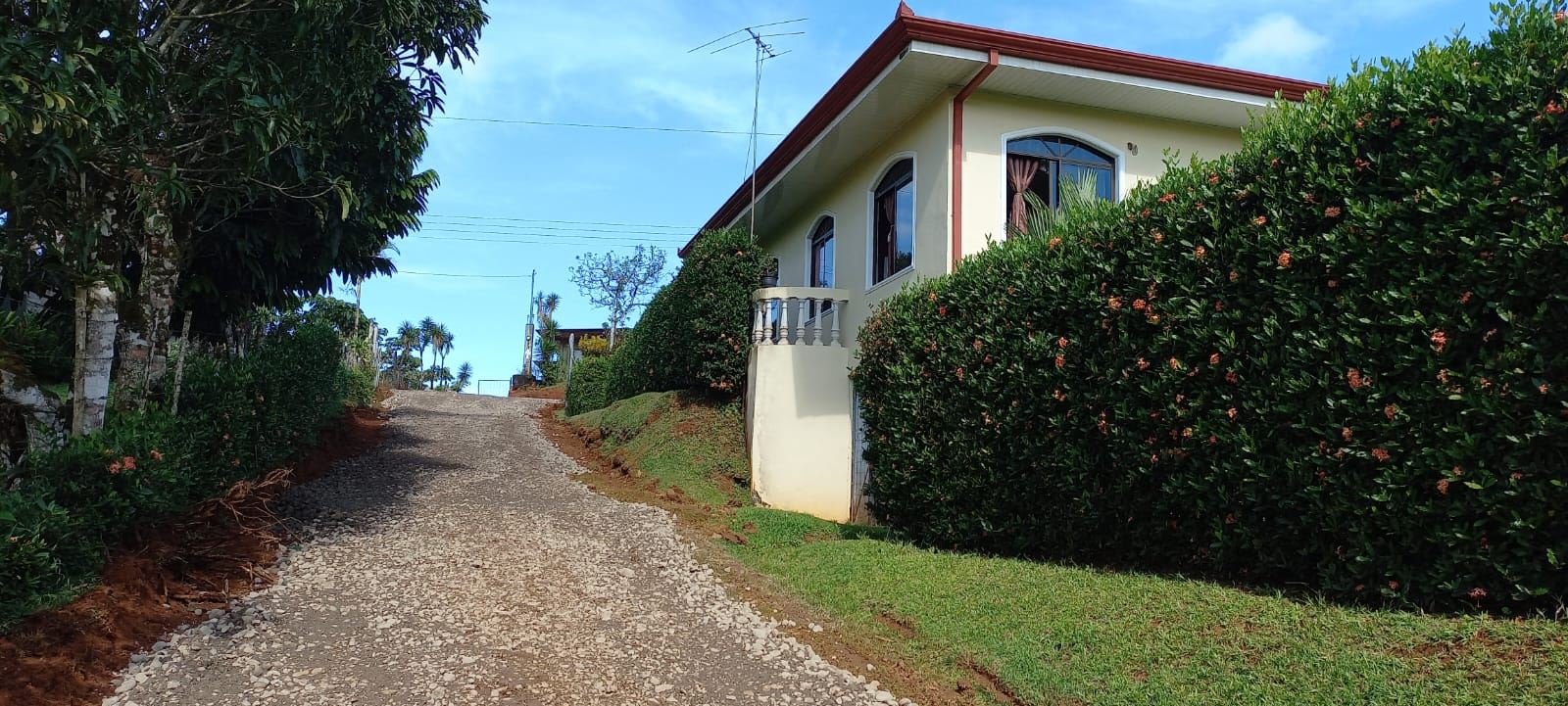 Gravel road slopes uphill beside a yellow house with arched windows, bordered by tall hedges under a blue sky.