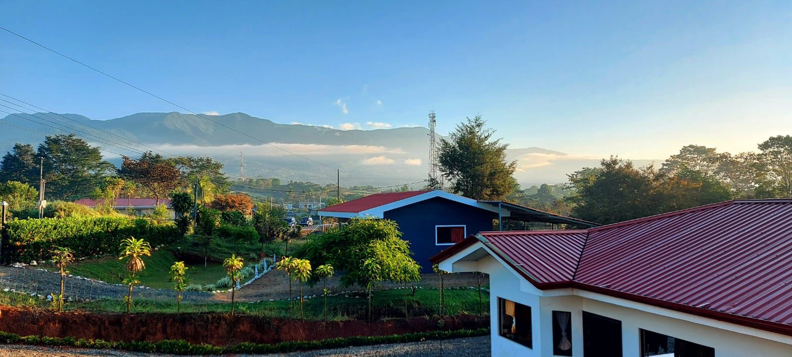 Rural neighborhood at dawn with red-roof houses, green gardens, and distant mountains under a blue sky.