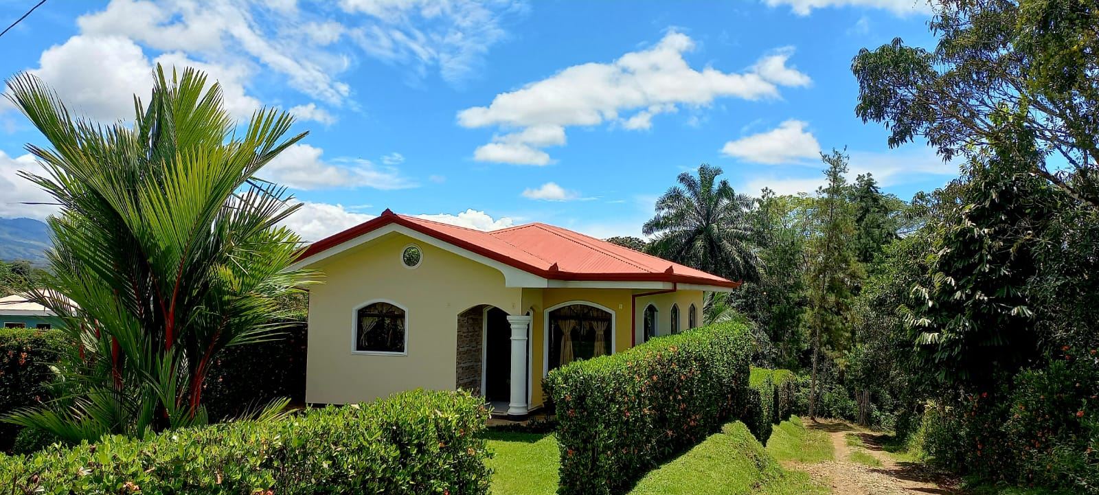Single-story yellow house with a red roof, arched windows, white columns, and a manicured hedge in a tropical garden on a sunny day