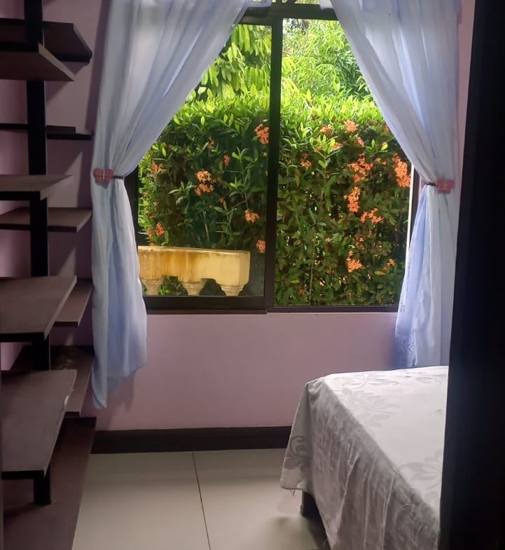Bedroom with a large window framed by light blue curtains, open to a garden view of green plants and orange flowers; a bed with a white patterned bedspread is on the right, and dark wooden shelves run along the left wall.