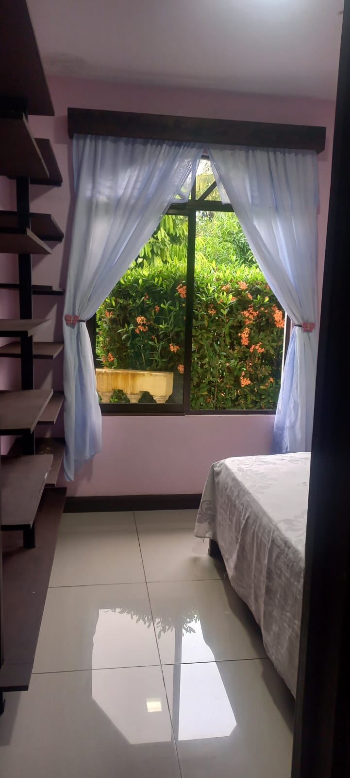 Bedroom with a large window framed by light blue curtains, open to a garden view of green plants and orange flowers; a bed with a white patterned bedspread is on the right, and dark wooden shelves run along the left wall.
