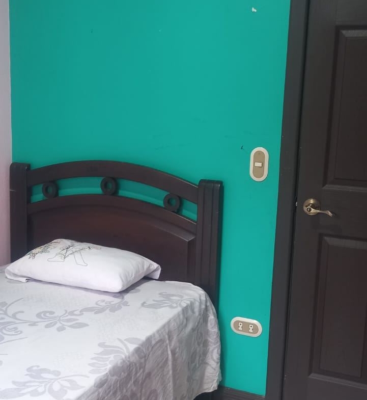 Teal bedroom wall with a dark wooden bed frame and a white floral bedspread, pillow, beside a dark door.