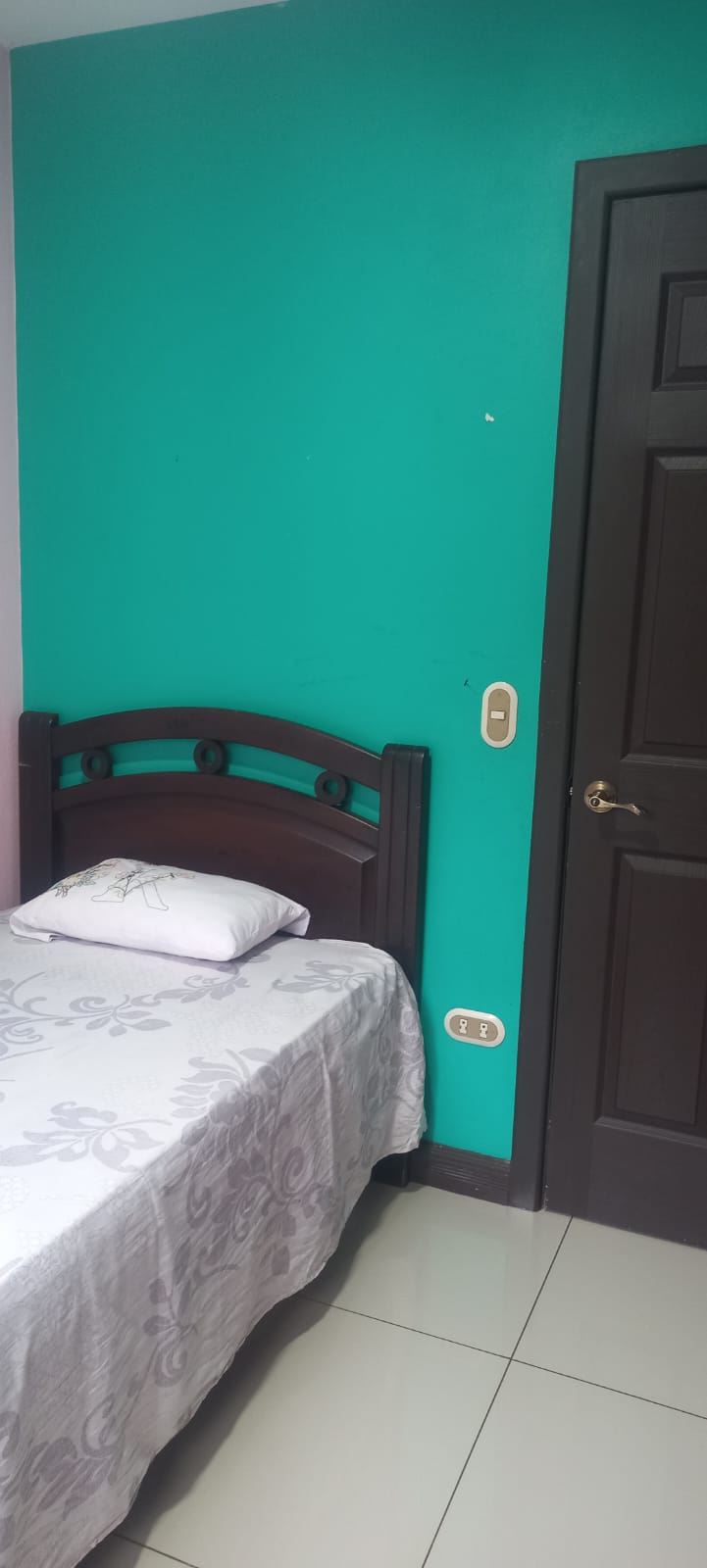 Teal bedroom wall with a dark wooden bed frame and a white floral bedspread, pillow, beside a dark door.