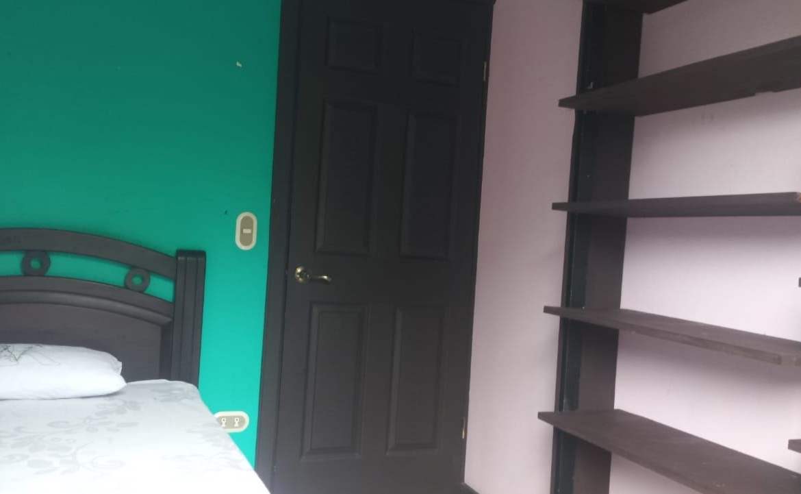 Bedroom with teal wall, black bed frame, and white patterned bedding next to a dark door and empty brown shelves on the right.