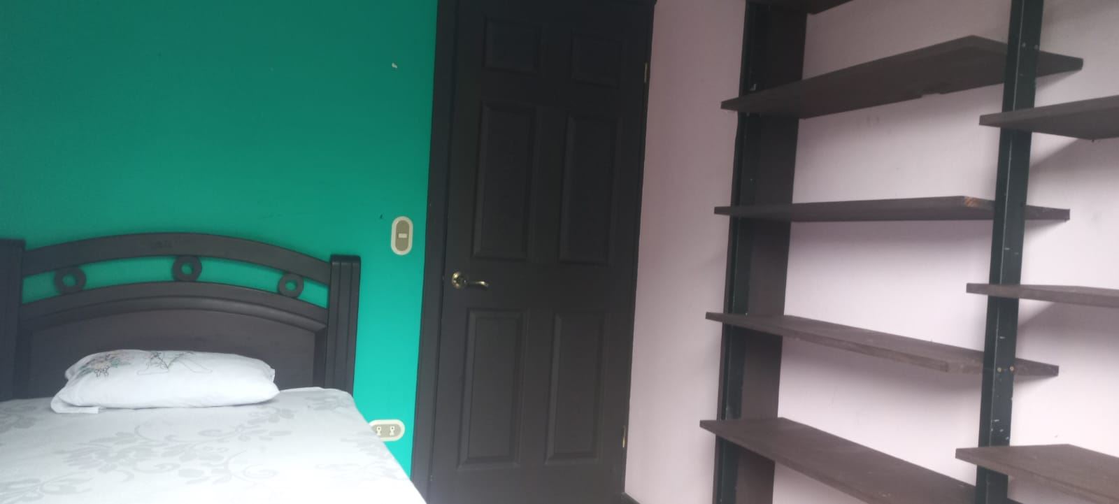 Bedroom with teal wall, black bed frame, and white patterned bedding next to a dark door and empty brown shelves on the right.
