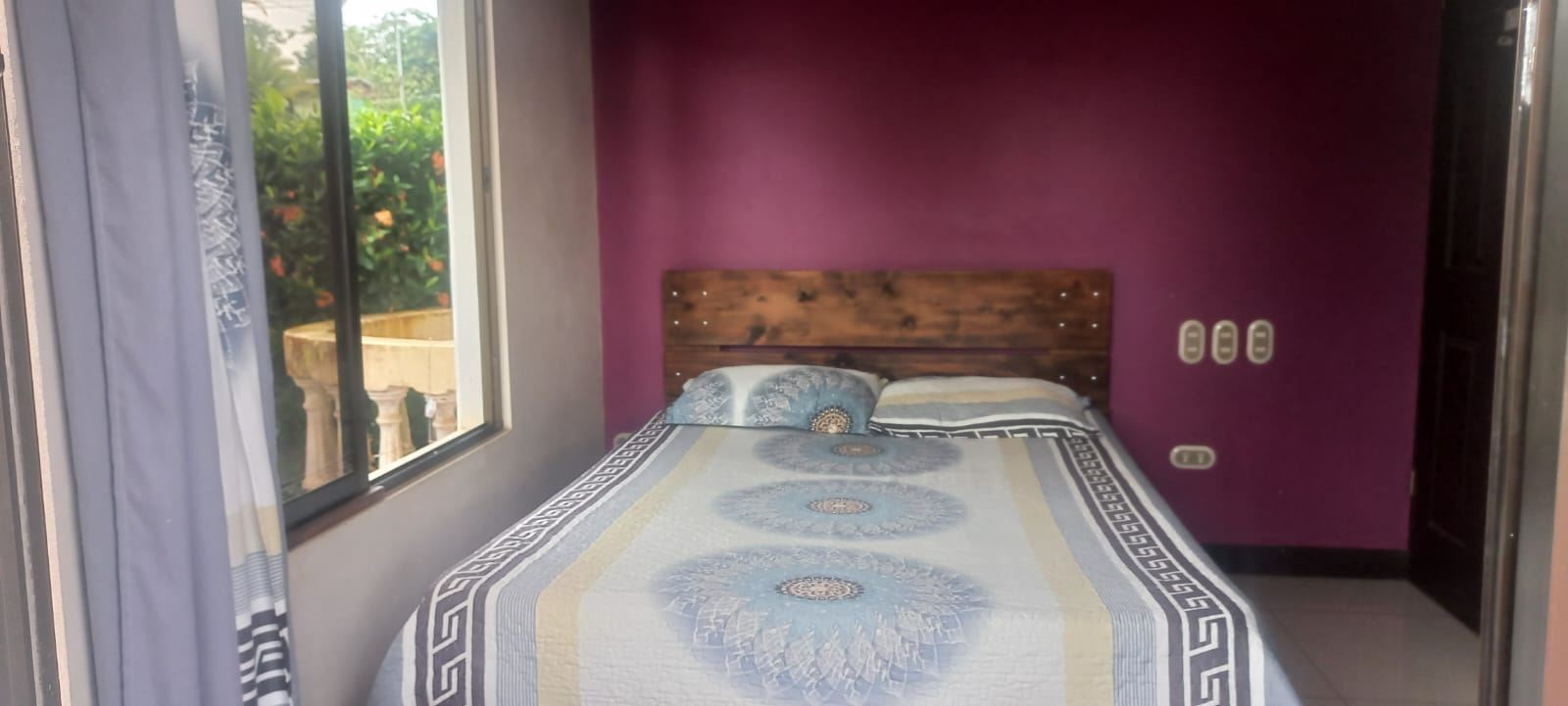 Bedroom with a wooden headboard bed against a purple accent wall, blue patterned quilt, and a window with curtains to the left.