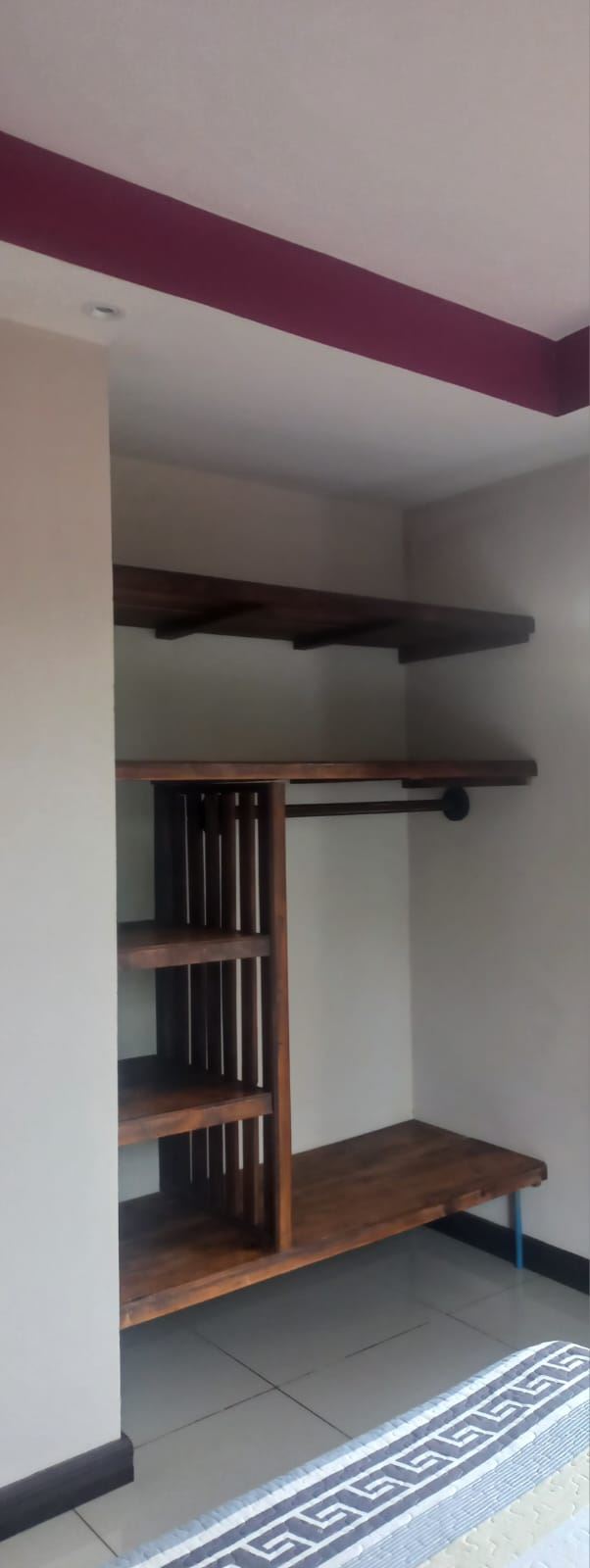Open wooden closet with vertical slatted divider, two shelves, a hanging rod, and a lower bench in a neutral-toned bedroom corner.