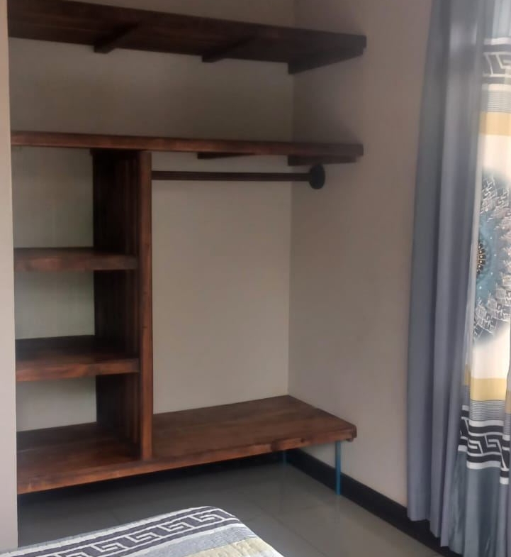 Corner bedroom with dark wood shelving and a built-in desk; a hanging rod for clothes on the right, and a bed with a blue patterned quilt in the foreground.