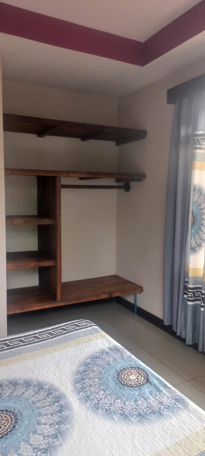 Corner bedroom with dark wood shelving and a built-in desk; a hanging rod for clothes on the right, and a bed with a blue patterned quilt in the foreground.