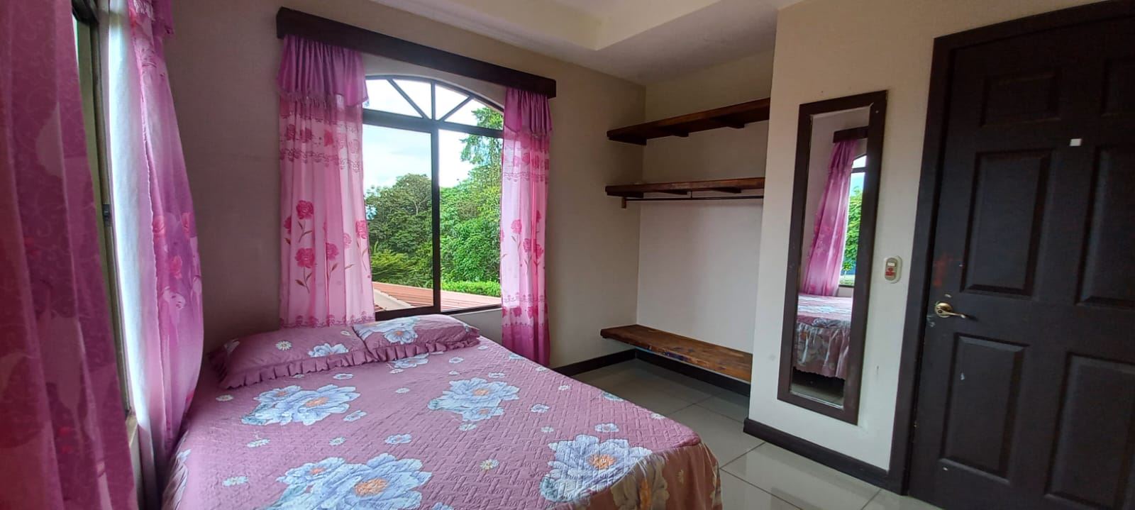 Bedroom with a pink floral bedspread, pink curtains, and a window showing green trees outside.