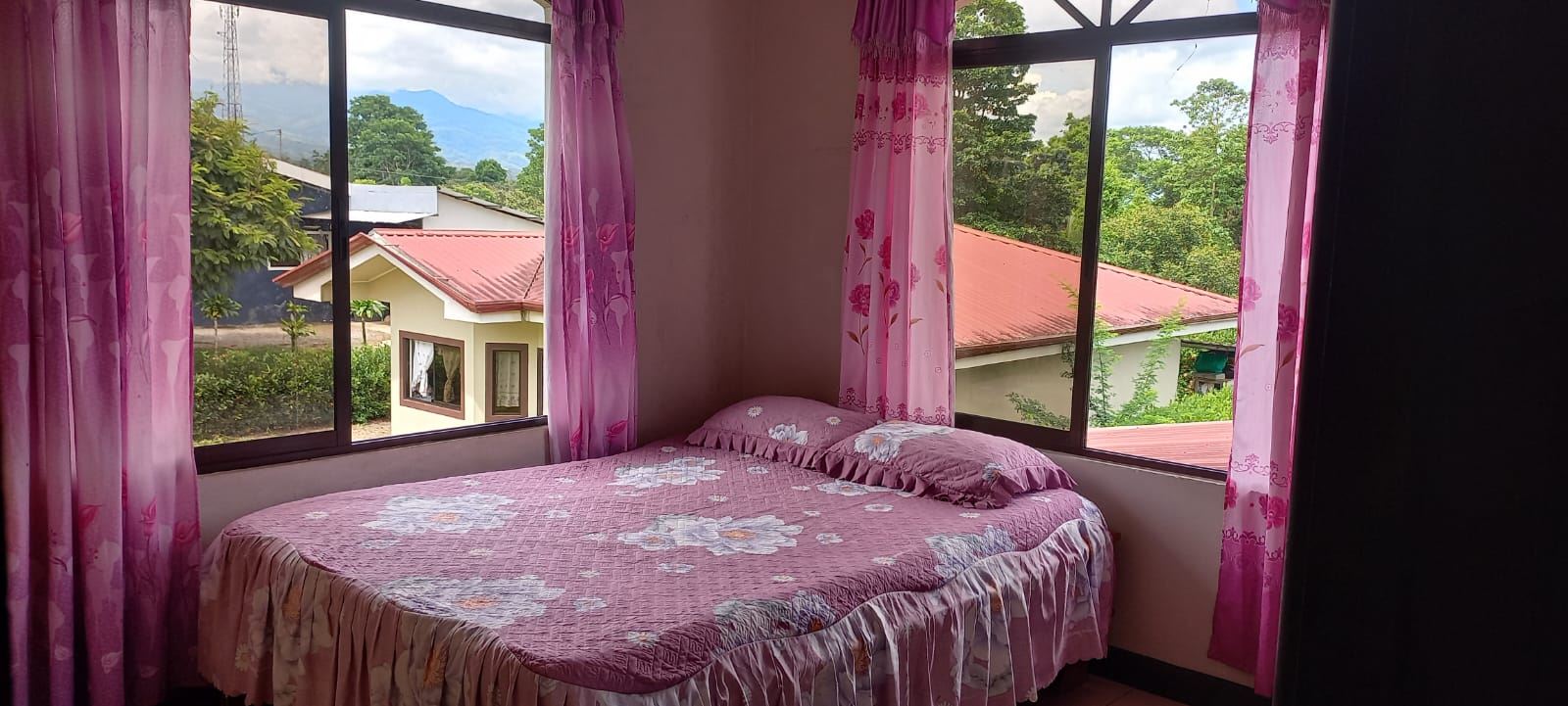 Corner bedroom with a pink floral bedspread and curtains, two pillows, and large windows overlooking red-roofed houses and green trees outside.