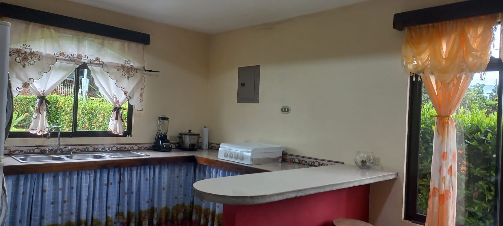 Cozy kitchen with a white countertop bar, pink base, and blue curtain-skirted cabinets beneath a double sink; window with decorative white curtains and greenery outside.
