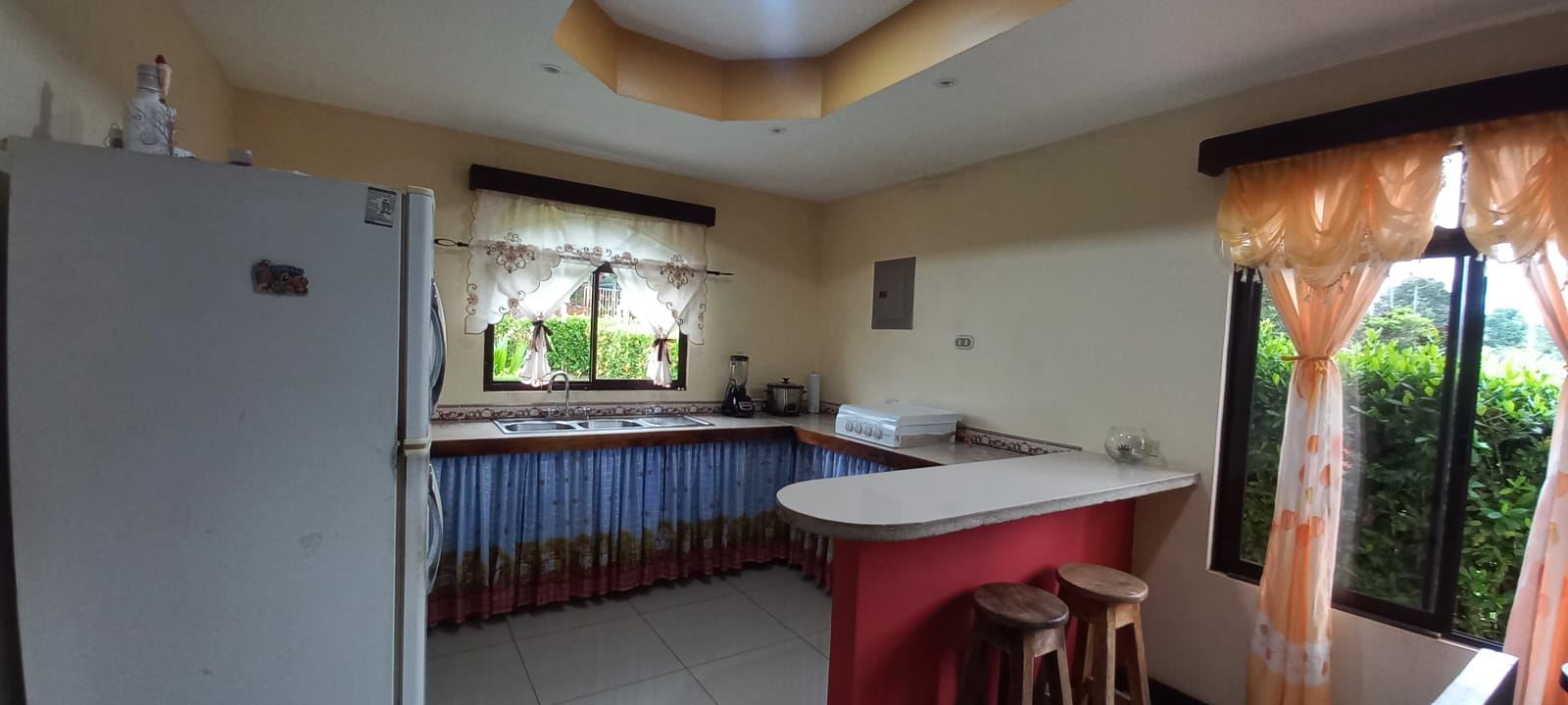 Kitchen with a white fridge on the left, dual sink under a window, blue ruffled cabinet skirts, and a red-bar island with two stools.