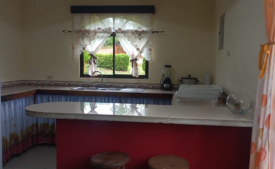 Kitchen with a white fridge on the left, sink under a window, and a red island with two wooden stools in the foreground.